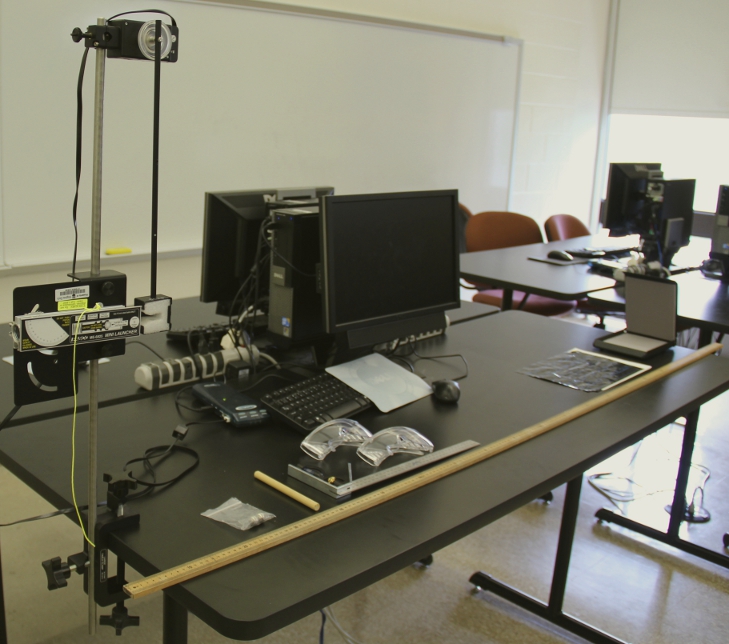 The full table set up of the ballistic pendulum lab. The launcher and pendulum are on the left. The projectile stop and paper are on the right. The loose items are between with the 2 meter stick across the table with 0 near the pendulum and the other end near the bin. 